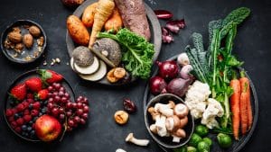 Eine Auswahl an frischem Obst und Gemüse, darunter Erdbeeren, Äpfel, Weintrauben, Karotten, Blumenkohl, Grünkohl, Champignons, Süßkartoffeln und Nüsse, stilvoll auf dunklen Tellern arrangiert. Die Zutaten präsentieren sich in leuchtenden Farben und symbolisieren die Vielfalt einer pflanzlichen Ernährung.