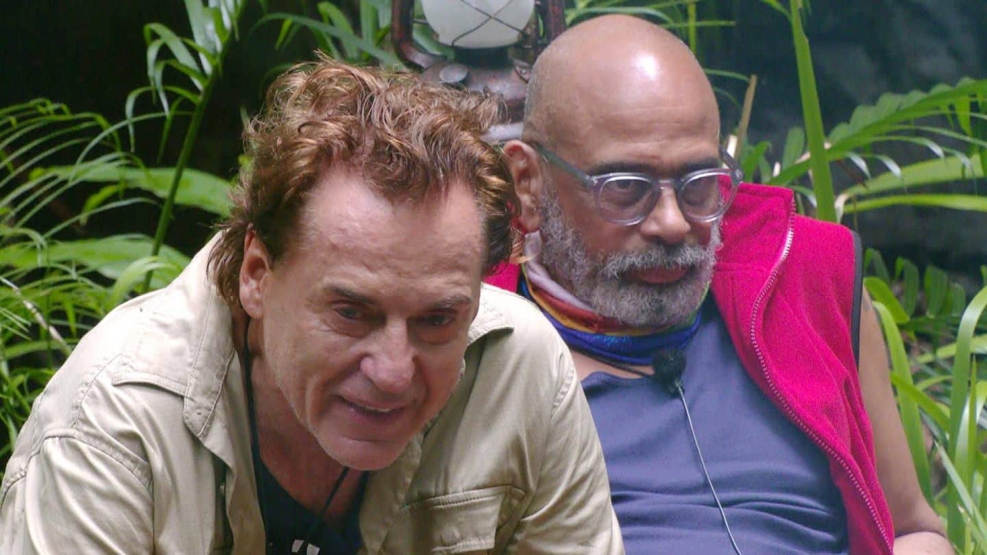 Jörg Dahlmann (links) und Pierre Sanoussi-Bliss (rechts) sitzen im Dschungelcamp nah beieinander und führen eine intensive Diskussion. Während Jörg eine nachdenkliche Miene zeigt, wirkt Pierre zurückhaltend. Die Szene spielt sich vor dichtem Dschungelgrün ab, eine Laterne hängt im Hintergrund.