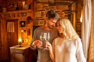 Ein verliebtes Paar stößt mit Sektgläsern in einer gemütlichen, rustikalen Holzhütte an. Die warme Beleuchtung unterstreicht die romantische Atmosphäre.