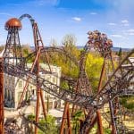 Die Achterbahn „Voltron Nevera“ im Europa-Park schlängelt sich mit ihren dunklen Schienen durch eine thematisch gestaltete Kulisse. Fahrgäste erleben eine steile Abfahrt, während sich die Strecke mit Loopings und Kurven durch die Landschaft windet. Im Hintergrund sind weitere Parkattraktionen und eine hügelige Landschaft zu sehen.