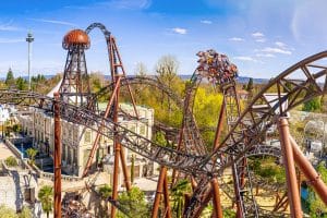 Die Achterbahn „Voltron Nevera“ im Europa-Park schlängelt sich mit ihren dunklen Schienen durch eine thematisch gestaltete Kulisse. Fahrgäste erleben eine steile Abfahrt, während sich die Strecke mit Loopings und Kurven durch die Landschaft windet. Im Hintergrund sind weitere Parkattraktionen und eine hügelige Landschaft zu sehen.