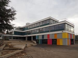 Das Friedrich-Schiller-Gymnasium in Marbach am Neckar. Das moderne Schulgebäude hat eine mehrstöckige Glas- und Fassadenkonstruktion mit bunten Farbakzenten an der unteren Etage. Eine breite Treppenanlage führt zum Haupteingang, während ein Baum im Vordergrund das Bild ergänzt. Der Himmel ist bewölkt.