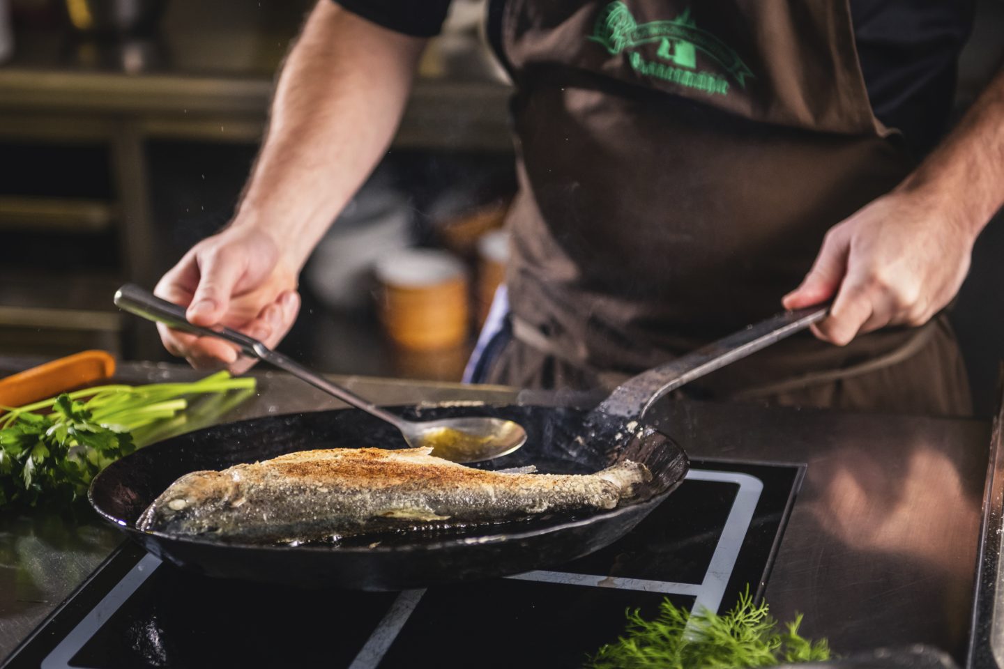 Ein Koch bereitet eine ganze Forelle in der Pfanne zu, während er sie mit heißer Butter übergießt. Frische Kräuter liegen auf der Arbeitsfläche.








