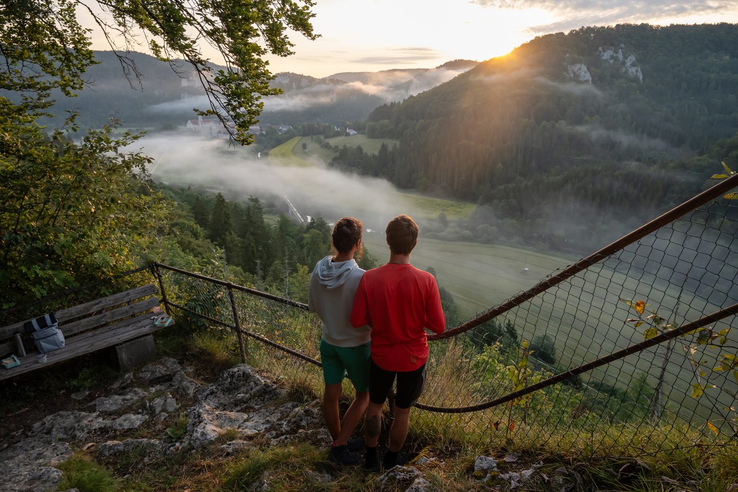 Zwei Menschen stehen auf einem Aussichtspunkt, blicken in ein von Nebel bedecktes, idyllisches Tal mit Wäldern, Feldern und Hügeln, während die Sonne am Horizont aufgeht und das Landschaftspanorama in warmes Licht taucht.