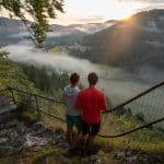Zwei Menschen stehen auf einem Aussichtspunkt, blicken in ein von Nebel bedecktes, idyllisches Tal mit Wäldern, Feldern und Hügeln, während die Sonne am Horizont aufgeht und das Landschaftspanorama in warmes Licht taucht.
