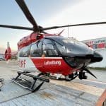 Roter und weißer Rettungshubschrauber der DRF Luftrettung, stationiert auf einem Landeplatz vor einem Gebäude mit der Aufschrift "DRF Luftrettung". Der Hubschrauber ist startklar und steht im Vordergrund, während der Himmel im Hintergrund leicht bewölkt ist.