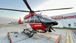 Roter und weißer Rettungshubschrauber der DRF Luftrettung, stationiert auf einem Landeplatz vor einem Gebäude mit der Aufschrift "DRF Luftrettung". Der Hubschrauber ist startklar und steht im Vordergrund, während der Himmel im Hintergrund leicht bewölkt ist.