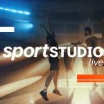 Ein Handballspiel in einer Halle mit dramatischer Beleuchtung. Ein Spieler in weißem Trikot springt mit dem Ball in der Hand, während ein Gegenspieler versucht, ihn zu blocken. Im Hintergrund steht ein Torwart vor dem Tor, bereit zur Abwehr. Das ZDF-Logo und der Schriftzug "sportSTUDIO live" sind über das Bild gelegt.