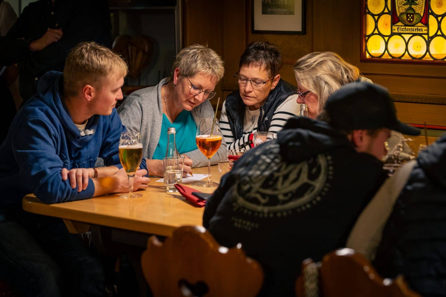 Eine Gruppe von vier Personen sitzt in einer Gaststube an einem Holztisch, schreibt konzentriert auf einen Zettel und löst zusammen ein Quiz.






