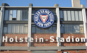 Außenansicht des Holstein-Stadions in Kiel mit dem Logo des Kieler SV Holstein von 1900 e.V. und dem Schriftzug 'Holstein-Stadion' auf der roten Ziegelfassade.
