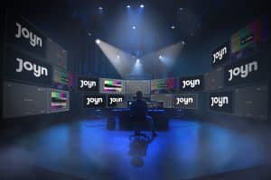 Ein Kontrollraum mit zahlreichen Bildschirmen, die das Joyn-Logo sowie Testbilder und "No Signal"-Hinweise zeigen. Im Zentrum sitzt eine Person vor einem Monitor und überwacht die Szene. Die düstere Beleuchtung und der Nebel erzeugen eine spannende Atmosphäre, passend zur neuen Staffel von "Big Brother"