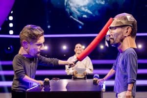 Joko Winterscheidt und Klaas Heufer-Umlauf tragen überdimensionale Masken und duellieren sich mit einem roten Schaumstoffstab, während Jeannine Michaelsen im Hintergrund moderiert. Die Szene spielt in einem bunten Studio mit futuristischer Beleuchtung.