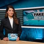 Linda Zervakis in einem schwarzen Blazer mit weißer Bluse steht hinter einem Studiotisch und hält eine Karte mit der Aufschrift „Fake News – Alles erstunken und erlogen“ in den Händen. Im Hintergrund ist ein großer Bildschirm mit dem gleichen Logo und einem stilisierten Globus zu sehen. Das Studio-Design ist in Blautönen gehalten, mit modernen grafischen Elementen.