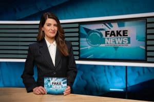 Linda Zervakis in einem schwarzen Blazer mit weißer Bluse steht hinter einem Studiotisch und hält eine Karte mit der Aufschrift „Fake News – Alles erstunken und erlogen“ in den Händen. Im Hintergrund ist ein großer Bildschirm mit dem gleichen Logo und einem stilisierten Globus zu sehen. Das Studio-Design ist in Blautönen gehalten, mit modernen grafischen Elementen.