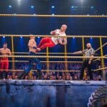 Fabian Hambüchen führt einen Dropkick gegen René Casselly im Wrestling-Ring aus, während ein Schiedsrichter und das Publikum die actionreiche Szene beobachten.