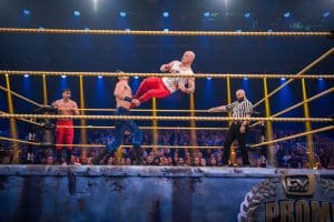 Fabian Hambüchen führt einen Dropkick gegen René Casselly im Wrestling-Ring aus, während ein Schiedsrichter und das Publikum die actionreiche Szene beobachten.