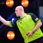 Michael van Gerwen, in grünem Trikot, konzentriert sich beim Wurf während der „Promi-Darts-WM“ 2024. Im Hintergrund leuchten orangefarbene Lampen, die die Bühne beleuchten.