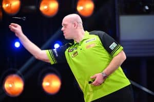 Michael van Gerwen, in grünem Trikot, konzentriert sich beim Wurf während der „Promi-Darts-WM“ 2024. Im Hintergrund leuchten orangefarbene Lampen, die die Bühne beleuchten.