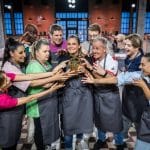 Die Teilnehmer der neuen Staffel von „Das große Promibacken“ posieren gemeinsam in einer Backstube. Sie tragen graue Schürzen und umringen den goldenen Cupcake-Pokal, den eine Teilnehmerin in den Händen hält. Die Backstube im Hintergrund ist modern eingerichtet mit beleuchteten Regalen und Küchenutensilien.