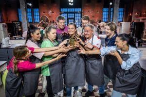 Die Teilnehmer der neuen Staffel von „Das große Promibacken“ posieren gemeinsam in einer Backstube. Sie tragen graue Schürzen und umringen den goldenen Cupcake-Pokal, den eine Teilnehmerin in den Händen hält. Die Backstube im Hintergrund ist modern eingerichtet mit beleuchteten Regalen und Küchenutensilien.