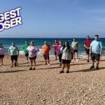 Eine Gruppe von 14 Teilnehmerinnen und Teilnehmern der Show „The Biggest Loser“ steht in Sportkleidung am Strand der Insel Naxos. Im Hintergrund sind das Meer und ein blauer Himmel zu sehen. Links oben ist das Logo von „The Biggest Loser“ eingeblendet. Die Teilnehmenden wirken motiviert und bereit für die Herausforderung.