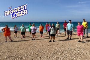 Eine Gruppe von 14 Teilnehmerinnen und Teilnehmern der Show „The Biggest Loser“ steht in Sportkleidung am Strand der Insel Naxos. Im Hintergrund sind das Meer und ein blauer Himmel zu sehen. Links oben ist das Logo von „The Biggest Loser“ eingeblendet. Die Teilnehmenden wirken motiviert und bereit für die Herausforderung.