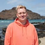 Knossi steht in einem orangefarbenen Hoodie am Strand, im Hintergrund türkisblaues Wasser, felsige Küsten und eine bergige Landschaft. Er blickt entspannt in die Kamera, die Sonne scheint, der Himmel ist leicht bewölkt.