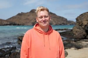 Knossi steht in einem orangefarbenen Hoodie am Strand, im Hintergrund türkisblaues Wasser, felsige Küsten und eine bergige Landschaft. Er blickt entspannt in die Kamera, die Sonne scheint, der Himmel ist leicht bewölkt.