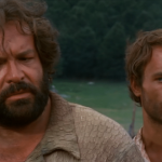 Szene aus einem Westernfilm mit zwei Männern in der Hauptrolle: Links ein kräftiger Mann mit Bart und lockigem Haar in einem beige-farbenen Hemd, rechts ein schlanker Mann mit kurzen braunen Haaren, leicht schmutzigem Gesicht und Hosenträgern. Im Hintergrund sind eine ländliche Szenerie mit Hütten, Kühen und Bäumen sowie eine weitere Person mit Hut zu sehen.