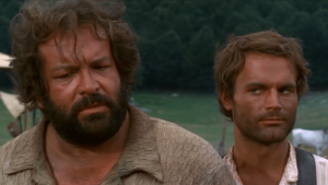 Szene aus einem Westernfilm mit zwei Männern in der Hauptrolle: Links ein kräftiger Mann mit Bart und lockigem Haar in einem beige-farbenen Hemd, rechts ein schlanker Mann mit kurzen braunen Haaren, leicht schmutzigem Gesicht und Hosenträgern. Im Hintergrund sind eine ländliche Szenerie mit Hütten, Kühen und Bäumen sowie eine weitere Person mit Hut zu sehen.