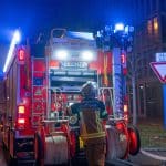 Ein Feuerwehrfahrzeug mit eingeschalteten Blaulichtern steht in der Nacht auf einer Straße. Ein Feuerwehrmann in voller Einsatzkleidung arbeitet an der Rückseite des Fahrzeugs, um Schläuche oder Ausrüstung vorzubereiten. Im Hintergrund sind Straßenschilder und ein Gebäude erkennbar.
