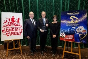 Drei Personen stehen vor einem grünen Vorhang. Links S.D. Fürst Albert II. von Monaco in dunklem Anzug mit Krawatte, in der Mitte Roland Mack, der eine Auszeichnung in den Händen hält, und rechts I.D. Prinzessin Stéphanie von Monaco in einem schwarzen Outfit. Links im Bild ein Aufsteller mit dem Logo der „Fédération Mondiale du Cirque“, rechts ein Plakat mit dem Europa-Park-Maskottchen und dem Text „50 – Célébrons sous les étoiles“.