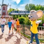 barrierefrei ChatGPT: ChatGPT Eine Familie mit zwei Kindern läuft fröhlich auf das Europa-Park-Maskottchen, eine große graue Maus in einem gelben T-Shirt mit blauem Stern, zu. Im Hintergrund sind eine Windmühle, eine Monorail-Bahn mit der Aufschrift „E.P. Express“ und blühende Blumenbeete zu sehen. Die Szene spielt sich auf einem gepflasterten Weg mit Geländer ab.