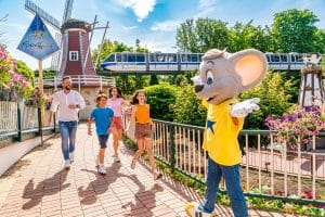 barrierefrei ChatGPT: ChatGPT Eine Familie mit zwei Kindern läuft fröhlich auf das Europa-Park-Maskottchen, eine große graue Maus in einem gelben T-Shirt mit blauem Stern, zu. Im Hintergrund sind eine Windmühle, eine Monorail-Bahn mit der Aufschrift „E.P. Express“ und blühende Blumenbeete zu sehen. Die Szene spielt sich auf einem gepflasterten Weg mit Geländer ab.