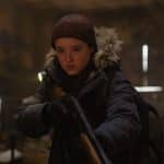 Bella Ramsey als Ellie in einer intensiven Szene aus der Serie "The Last of Us": Ellie trägt eine dicke Winterjacke mit Fellbesatz und eine Mütze. Sie hält ein Gewehr in den Händen und blickt konzentriert nach vorne. Die Umgebung ist dunkel und wirkt wie ein verlassenes Gebäude, das von spärlichem Licht durchbrochen wird.