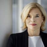 Porträtfoto von Julia Klöckner, die mit einem dezenten Lächeln in die Kamera blickt. Sie trägt einen schwarzen Blazer über einer weißen Bluse, ihre blonden Haare sind schulterlang gestylt. Der Hintergrund ist dezent verschwommen, mit einer hellen Wand auf der linken Seite und einem modernen Büroambiente auf der rechten.
