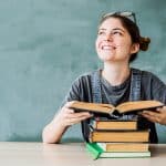 Eine junge Frau sitzt vor einer Tafel und hält ein aufgeschlagenes Buch in den Händen. Sie lächelt nach oben, während mehrere weitere Bücher auf dem Tisch gestapelt sind. Sie trägt eine graue Latzhose und hat ihre Brille auf den Kopf geschoben.