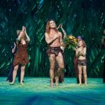 Alexander Klaws steht als Tarzan auf der Bühne des Stage Palladium Theaters in Stuttgart und erhält unter großem Applaus einen Blumenstrauß. Um ihn herum stehen Darstellerinnen und Darsteller in Dschungel-Kostümen, die ebenfalls klatschen und lächeln. Der Hintergrund ist in grünen Farbtönen gestaltet, passend zur Dschungel-Thematik des Musicals.