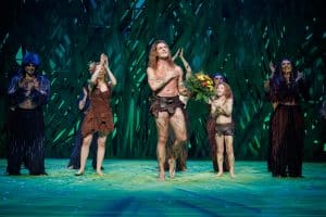 Alexander Klaws steht als Tarzan auf der Bühne des Stage Palladium Theaters in Stuttgart und erhält unter großem Applaus einen Blumenstrauß. Um ihn herum stehen Darstellerinnen und Darsteller in Dschungel-Kostümen, die ebenfalls klatschen und lächeln. Der Hintergrund ist in grünen Farbtönen gestaltet, passend zur Dschungel-Thematik des Musicals.