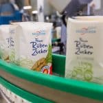 Packungen von „Feiner Rüben Zucker“ der Südzucker AG auf einem grünen Förderband in einer Produktionshalle. Im Hintergrund sind Maschinen und unscharfe Details der Produktion zu sehen.