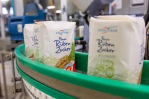 Packungen von „Feiner Rüben Zucker“ der Südzucker AG auf einem grünen Förderband in einer Produktionshalle. Im Hintergrund sind Maschinen und unscharfe Details der Produktion zu sehen.