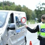 Eine Beamtin der Bundespolizei kontrolliert ein Fahrzeug an einer Grenzkontrolle. Sie hält ein rotes Halt-Schild mit der Aufschrift „HALT BUNDESPOLIZEI“ in Richtung des Fahrers. Der silberne Transporter wird an einem Kontrollpunkt mit Verkehrshütchen angehalten. Im Hintergrund sind Bäume und eine Straße zu sehen.