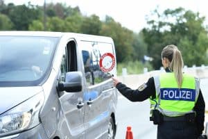 Eine Beamtin der Bundespolizei kontrolliert ein Fahrzeug an einer Grenzkontrolle. Sie hält ein rotes Halt-Schild mit der Aufschrift „HALT BUNDESPOLIZEI“ in Richtung des Fahrers. Der silberne Transporter wird an einem Kontrollpunkt mit Verkehrshütchen angehalten. Im Hintergrund sind Bäume und eine Straße zu sehen.