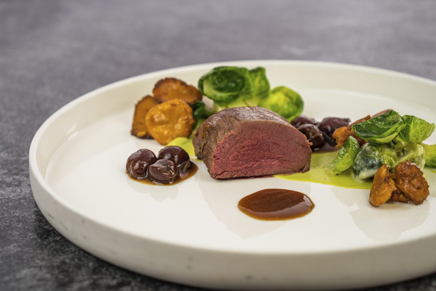 Gourmet-Wildgericht auf einem weißen Teller, serviert mit gebratenen Pilzen, Rosenkohl, dunkler Sauce und Maronen.







