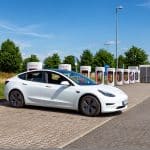 Ein weißer Tesla Model 3 steht an einer Tesla-Supercharger-Station auf einem Parkplatz bei sonnigem Wetter. Im Hintergrund sind mehrere Ladepunkte und eine grüne Landschaft mit Bäumen zu sehen.