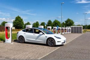 Ein weißer Tesla Model 3 steht an einer Tesla-Supercharger-Station auf einem Parkplatz bei sonnigem Wetter. Im Hintergrund sind mehrere Ladepunkte und eine grüne Landschaft mit Bäumen zu sehen.
