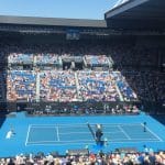die Rod Laver Arena im Melbourne Park während der Australian Open 2020. Es handelt sich um ein Viertelfinalspiel im Herreneinzel zwischen Alexander Zverev und Stan Wawrinka. Die Arena ist gut gefüllt mit Zuschauern, viele tragen Hüte und Sonnenbrillen. Der Tennisplatz ist in einem auffälligen Blauton gehalten, umgeben von Werbebannern, unter anderem von „KIA“ und „Emirates“. Spieler und Ballkinder sind auf dem Spielfeld verteilt, während sich das Publikum auf die Partie konzentriert. Ein Teil der Tribünen liegt im Schatten, während andere Bereiche von der Sonne beleuchtet werden.