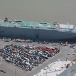 Eine Luftaufnahme eines Hafens, in dem mehrere Frachtschiffe festgemacht sind, darunter ein großes Autotransportschiff. Im Vordergrund sind zahlreiche parkende Autos und Lastwagen auf dem Hafengelände zu sehen. Der Hafen grenzt an einen breiten Fluss, mit klaren Straßen und Umschlagsbereichen für Fahrzeuge und Container.