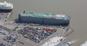 Eine Luftaufnahme eines Hafens, in dem mehrere Frachtschiffe festgemacht sind, darunter ein großes Autotransportschiff. Im Vordergrund sind zahlreiche parkende Autos und Lastwagen auf dem Hafengelände zu sehen. Der Hafen grenzt an einen breiten Fluss, mit klaren Straßen und Umschlagsbereichen für Fahrzeuge und Container.