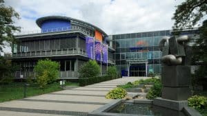 Das Hauptgebäude der badenova mit moderner Glasfassade und farbigen Akzenten, umgeben von einer gepflegten Gartenanlage. Im Vordergrund steht eine markante Skulptur mit integriertem Brunnen